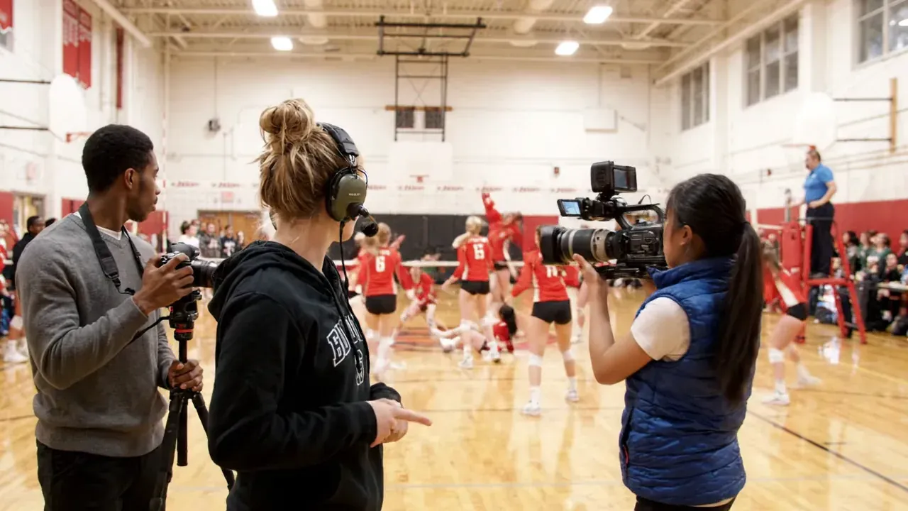 AI-generated scene of two student camera operators and a student director with a headset filming a high school volleyball match from the side of the gym floor
