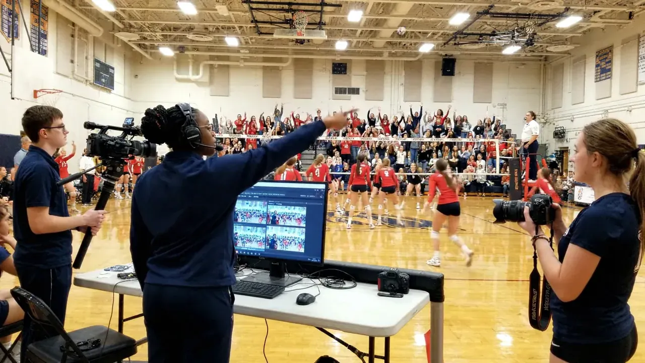 AI-generated scene of a student crew at a high school volleyball match: camera operators on the gym floor and a director pointing at monitors in a control position